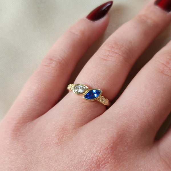 Gold ring with a a sapphire marquise and a pear cut diamond on a person's finger against a neutral background