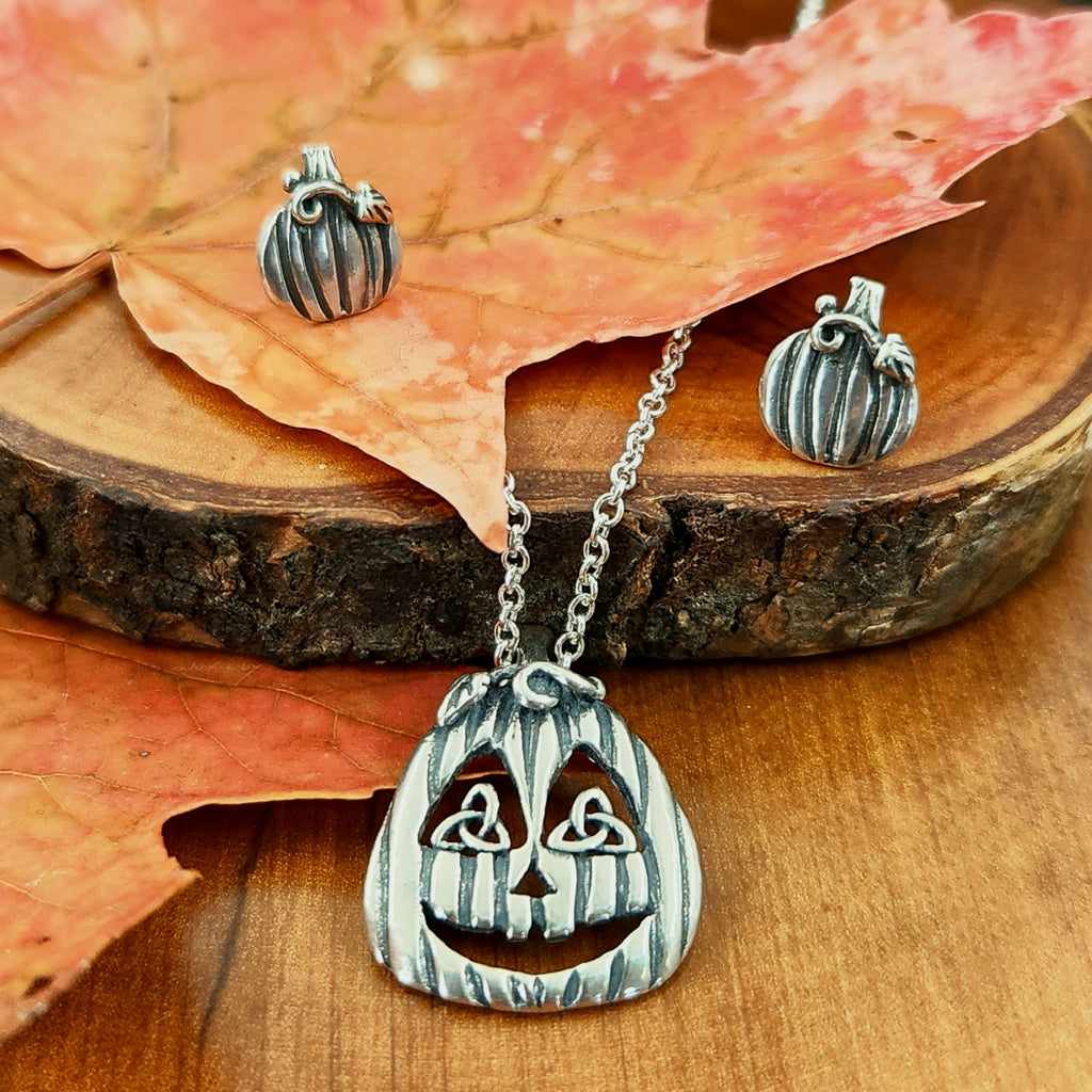 Pumpkin-shaped earrings and a jack-o'-lantern pendant on a wooden surface with fall leaves.