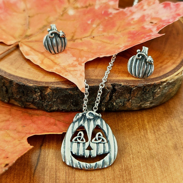 Pumpkin-shaped earrings and a jack-o'-lantern pendant on a wooden surface with fall leaves.