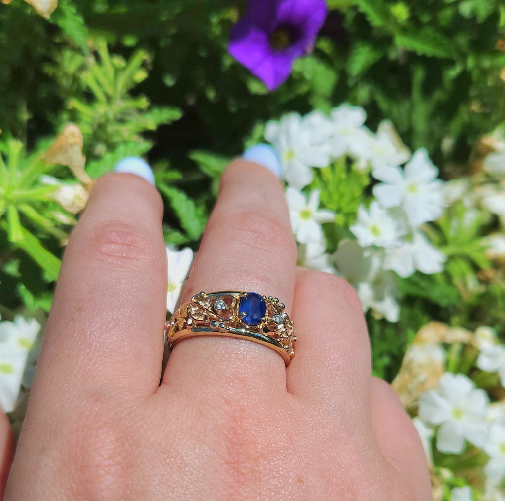 Gold custom frame ring with rose gold, white gold, and yellow gold set with two diamonds and an oval blue sapphire, shown on a hand surrounded b foliage.