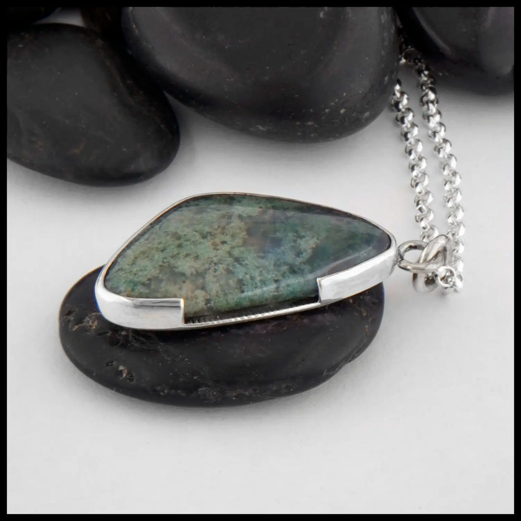 This custom Scalene Triangle Cabochon Moss Agate pendant is set in a custom open back, asymmetrical Sterling Silver Bezel. The backside, decorated with a 14 Karat Yellow Gold climbing spiral with the edges of the bezel. This photo includes a side shot of the pendant to show off the asymmetrical bezel with the break in the the frame to introduce a fork like pattern. Includes an 18 inch Medium Rolo Chain.