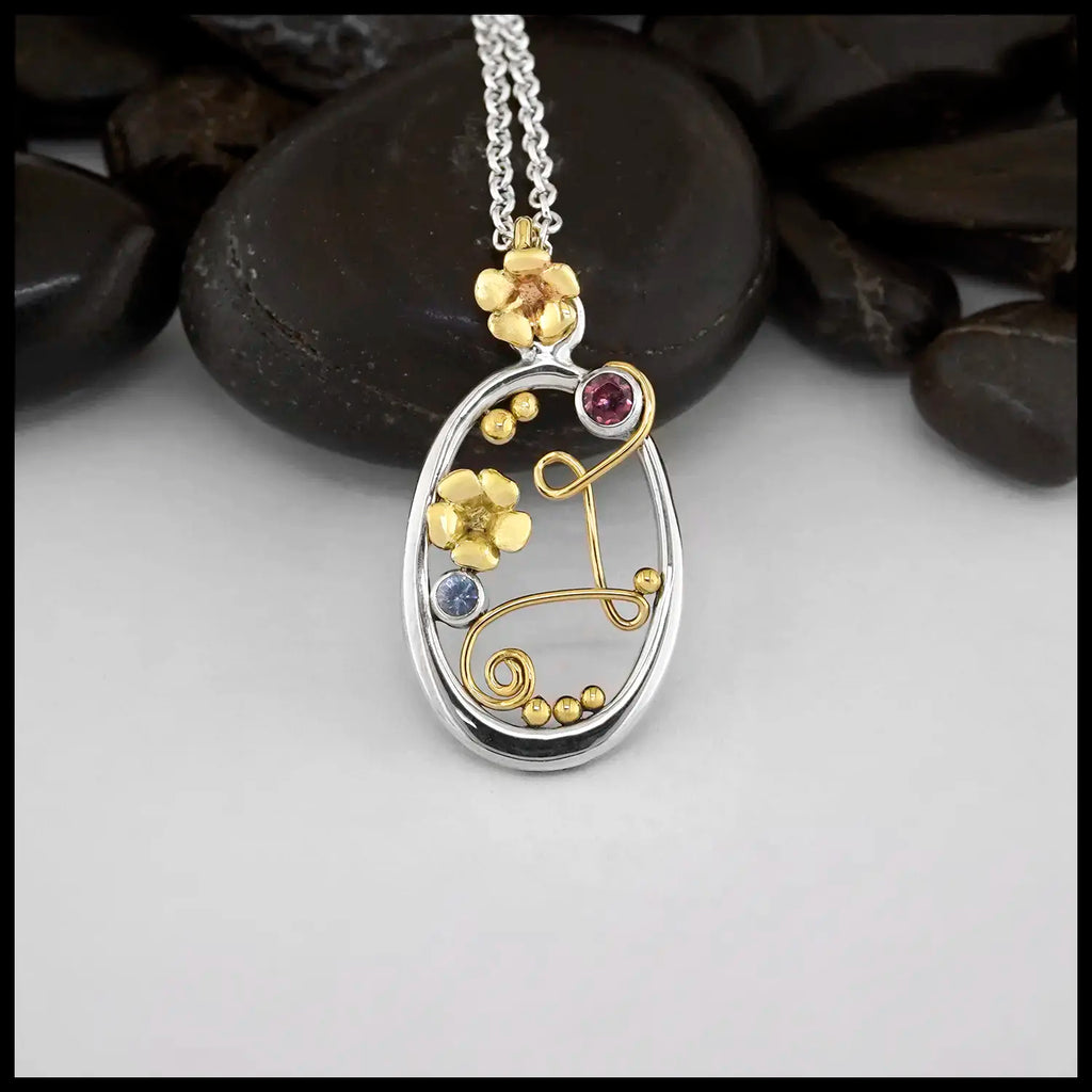Sterling Silver Oval Frame Pendant with a 14 Karat Yellow Gold Bryn's Blossom flower bale. Interrupting the frame on the top left is a bezel set Pink Tourmaline. Caressing the Tourmaline and making two loops across the frame to swirl at the bottom is an 14 Karat Yellow Gold Wire Swirl. Another 14K Yellow Gold Bryn's Blossom is set in the middle, accompanied by a Ceylon Sapphire. Yellow Gold Beading accenting the inside of the frame. Includes an 18" Cable Chain. Made by our Craftsman Sydney.