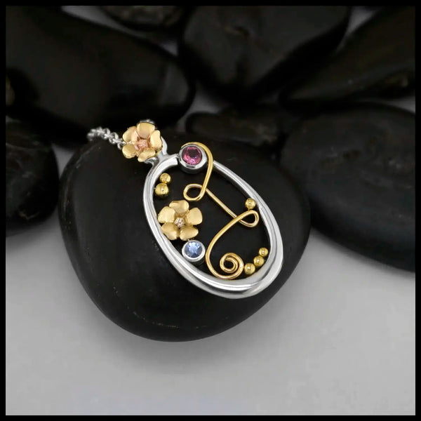 Sterling Silver Oval Frame Pendant with a 14 Karat Yellow Gold Bryn's Blossom flower bale. Interrupting the frame on the top left is a bezel set Pink Tourmaline. Caressing the Tourmaline and making two loops across the frame to swirl at the bottom is an 14 Karat Yellow Gold Wire Swirl. Another 14K Yellow Gold Bryn's Blossom is set in the middle, accompanied by a Ceylon Sapphire. Yellow Gold Beading accenting the inside of the frame. Includes an 18" Cable Chain. Made by our Craftsman Sydney.
