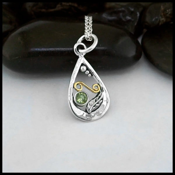 A small Tear Drop Frame in Sterling Silver. In the lower left is a Sterling Silver Feather charm, adjacent to the feather is a bezel set Peridot. Immediately above lies an 18 Karat Yellow Gold Spiral. Lastly there are three descending Sterling Silver beads on the inner top left of the frame. This pendant includes an 18" Cable Chain. Made by Sydney.