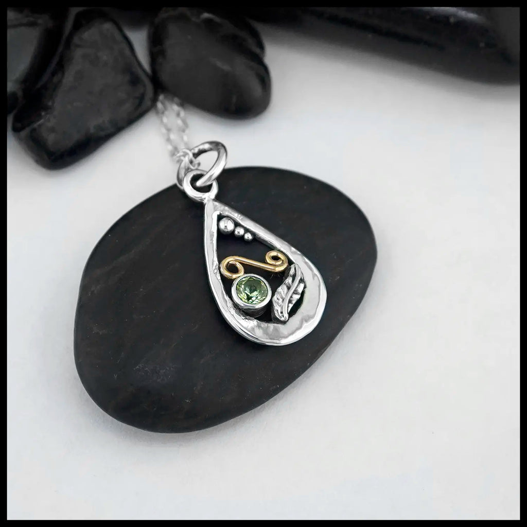 A small Tear Drop Frame in Sterling Silver. In the lower left is a Sterling Silver Feather charm, adjacent to the feather is a bezel set Peridot. Immediately above lies an 18 Karat Yellow Gold Spiral. Lastly there are three descending Sterling Silver beads on the inner top left of the frame. This pendant includes an 18" Cable Chain. Made by Sydney.