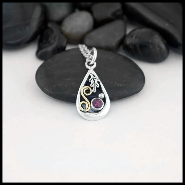 A small Tear Drop Frame in Sterling Silver. In the upper left is a Sterling Silver Oak leaf charm, below the Oak Leaf is a bezel set pink Tourmaline and a Sterling Silver beads. Filling the right side of the frame is an 18 Karat Yellow Gold Spiral. This pendant includes an 18" Cable Chain. Made by Sydney.