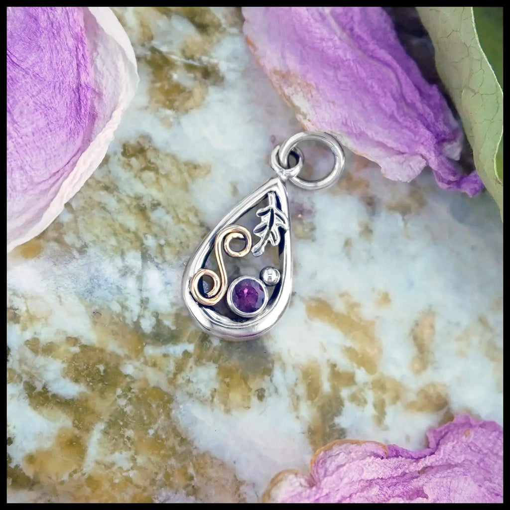 Upon Peony Petals and a slab of Connemara Marble, a small Tear Drop Frame in Sterling Silver. In the upper left is a Sterling Silver Oak leaf charm, below the Oak Leaf is a bezel set pink Tourmaline and a Sterling Silver beads. Filling the right side of the frame is an 18 Karat Yellow Gold Spiral. This pendant includes an 18" Cable Chain. Made by Sydney.