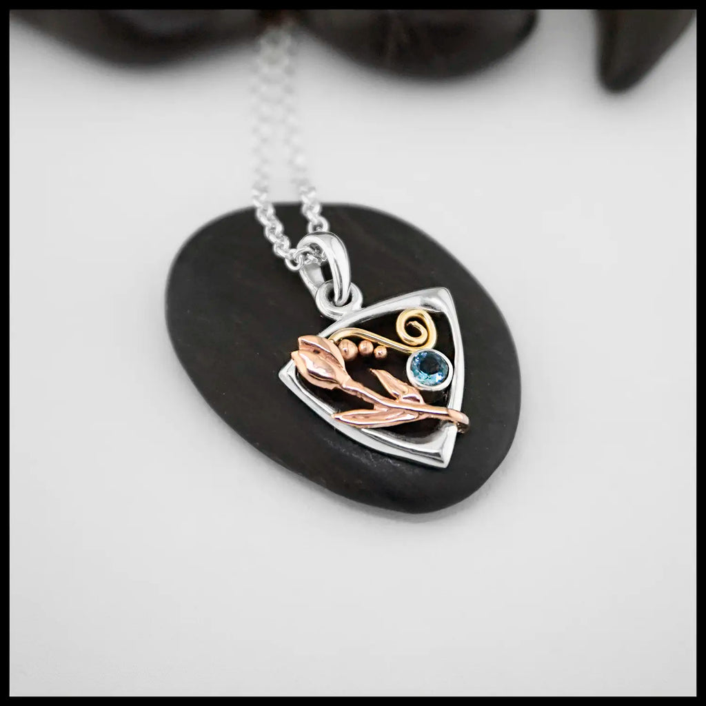 The latest necklace from out Craftsman Nicole! A Sterling Silver Triangular Frame Pendant with a 14K Yellow Gold Swirl accenting the top, a 14K Rose Gold Tulip & beading on the right side adjacent to a bezel set Aquamarine on the left. Includes an 18" Cable Chain 