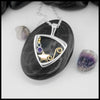 Triangular frame pendant with star and swirl design, with blue and purple stone settings. Laid on a black stone with scattered stones around the white background.