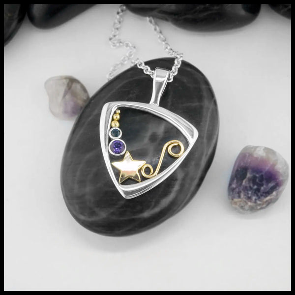 Triangular frame pendant with star and swirl design, with blue and purple stone settings. Laid on a black stone with scattered stones around the white background.