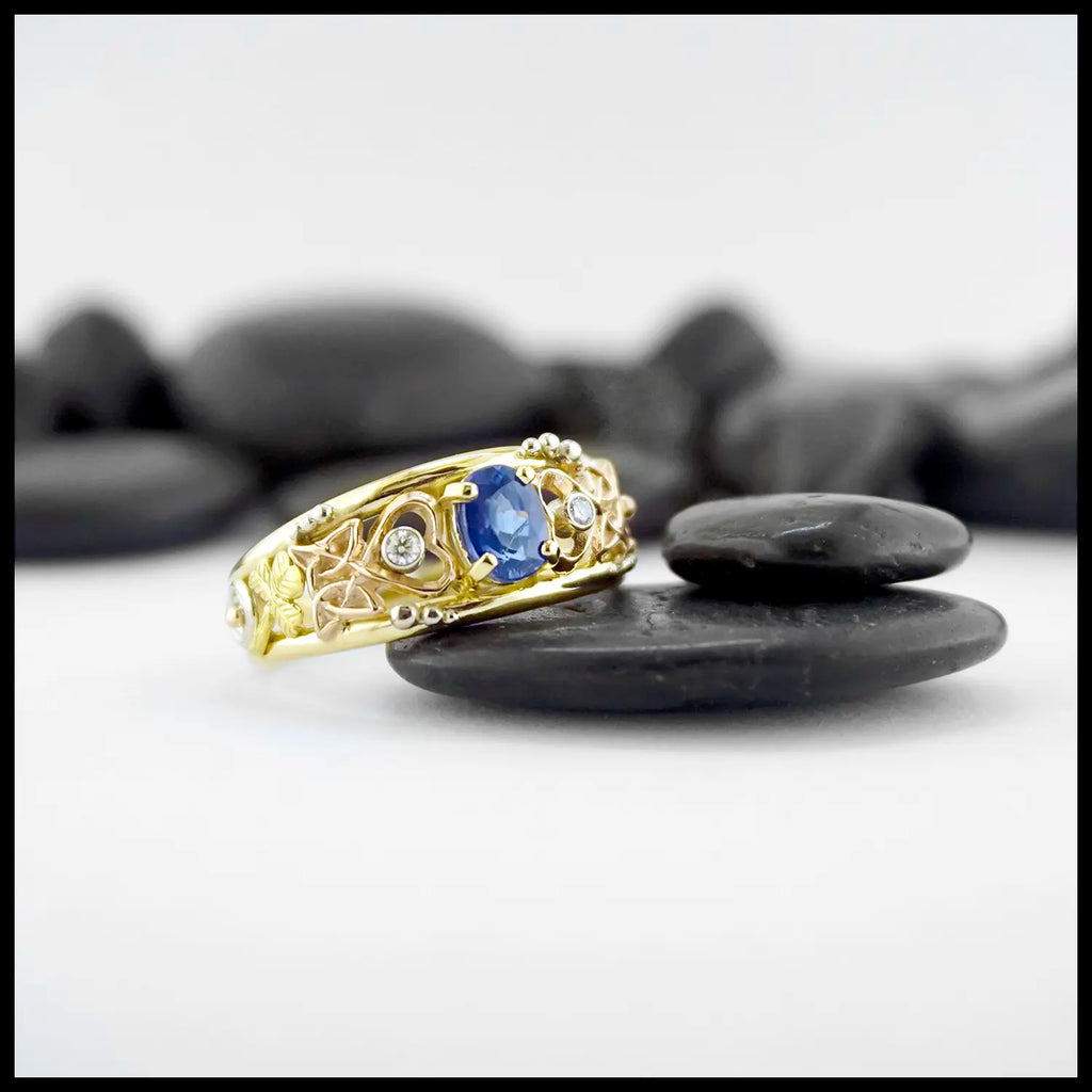 Gold custom frame ring with rose gold, white gold, and yellow gold set with two diamonds and an oval blue sapphire