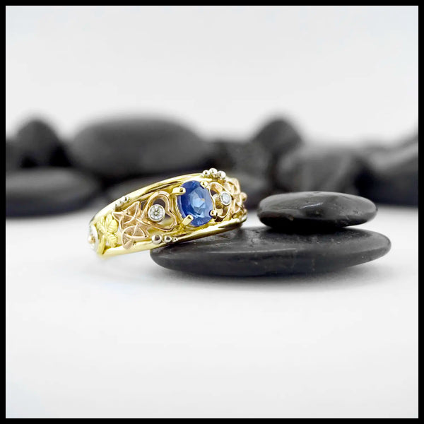 Gold custom frame ring with rose gold, white gold, and yellow gold set with two diamonds and an oval blue sapphire