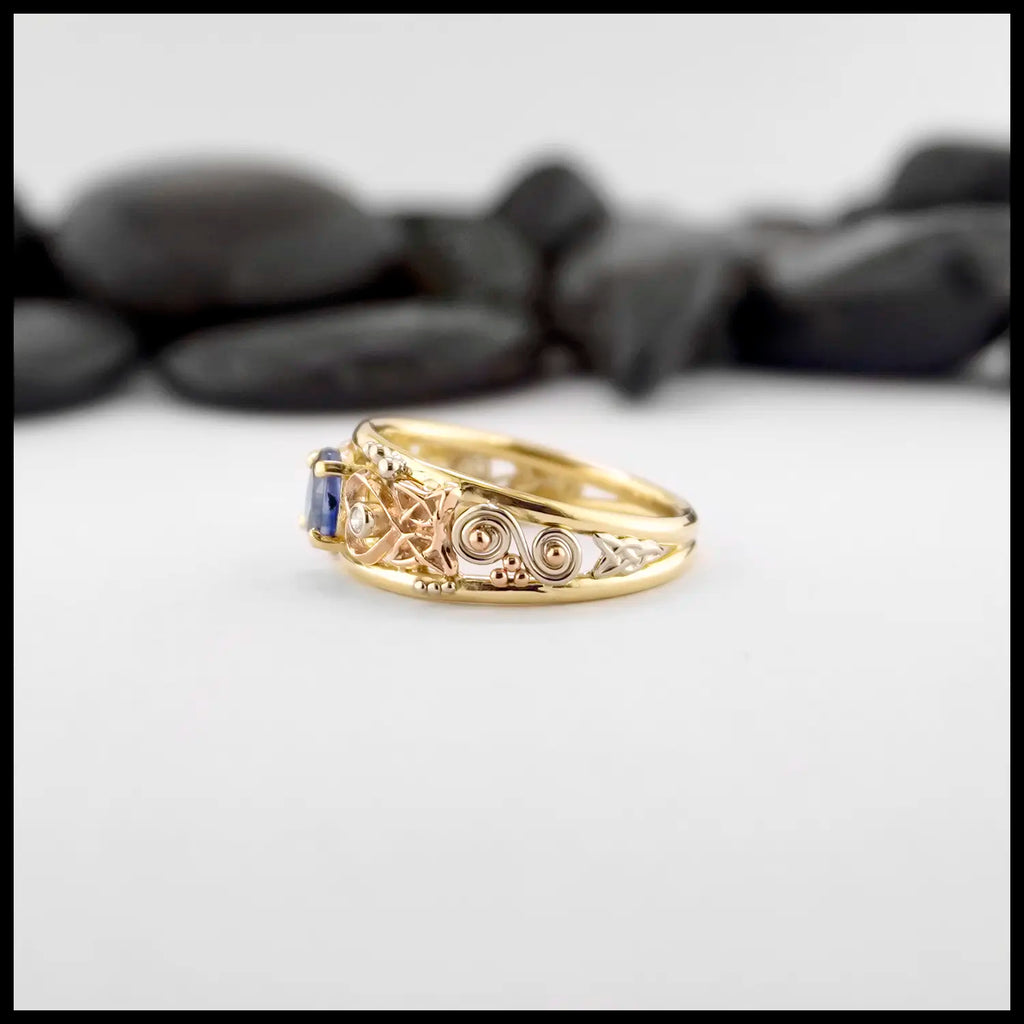 Gold custom frame ring with rose gold, white gold, and yellow gold set with two diamonds and an oval blue sapphire