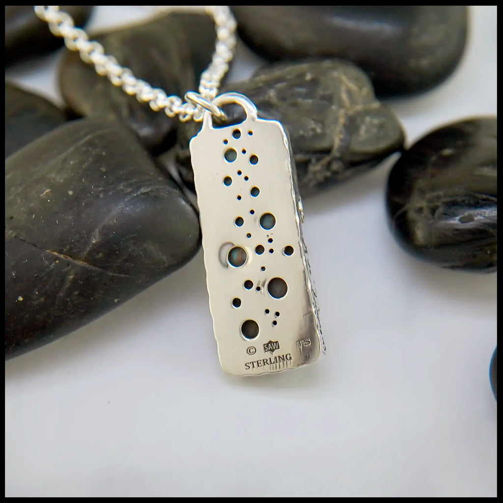 Silver pendant with engraved design on a chain, set against a background of dark stones.