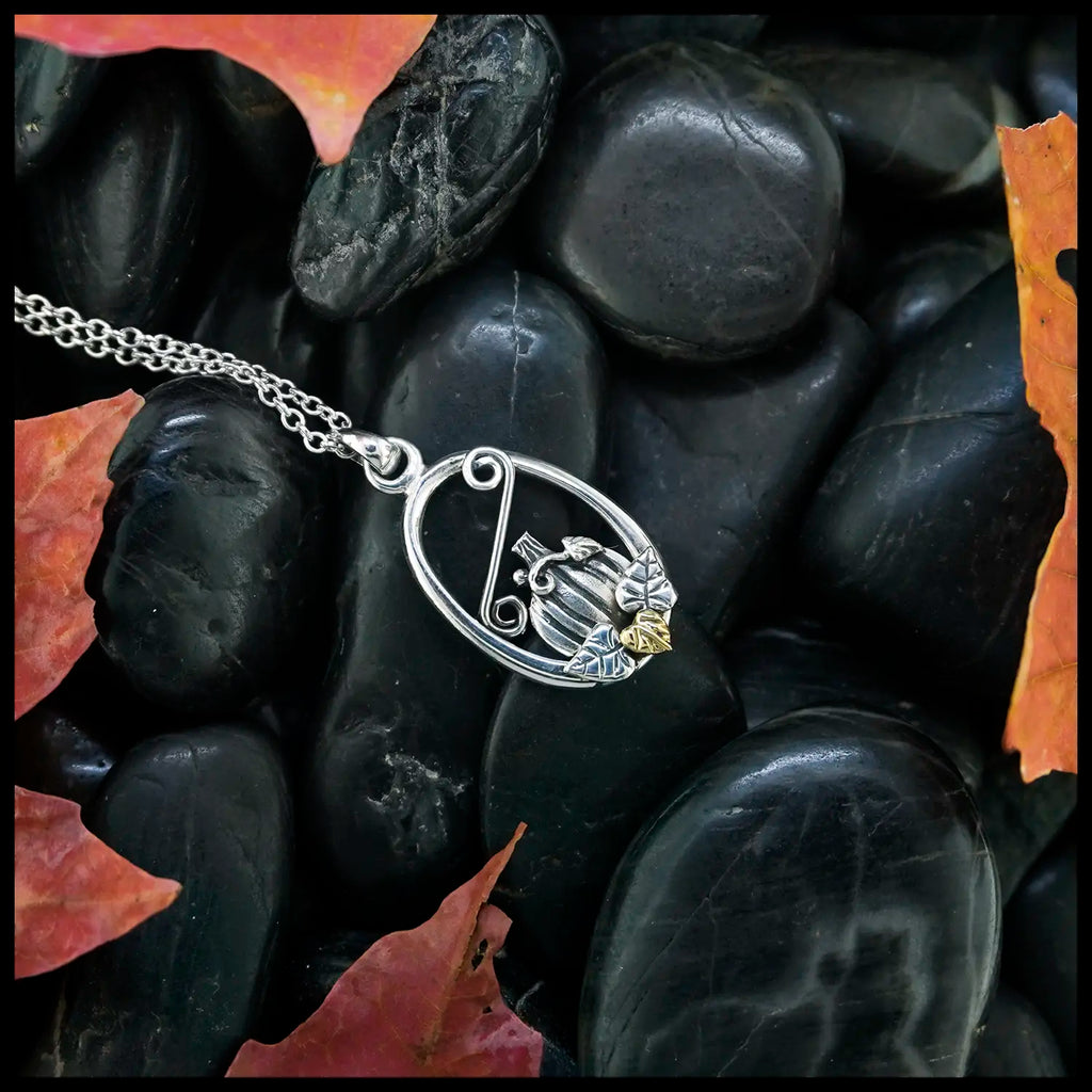 Silver pumpkin frame pendant with a 14K yellow gold leaf, on a chain, lying on black stones with autumn leaves.