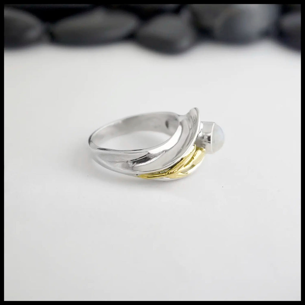 Silver and gold Moonstone ring on a white surface with blurred stones in the background