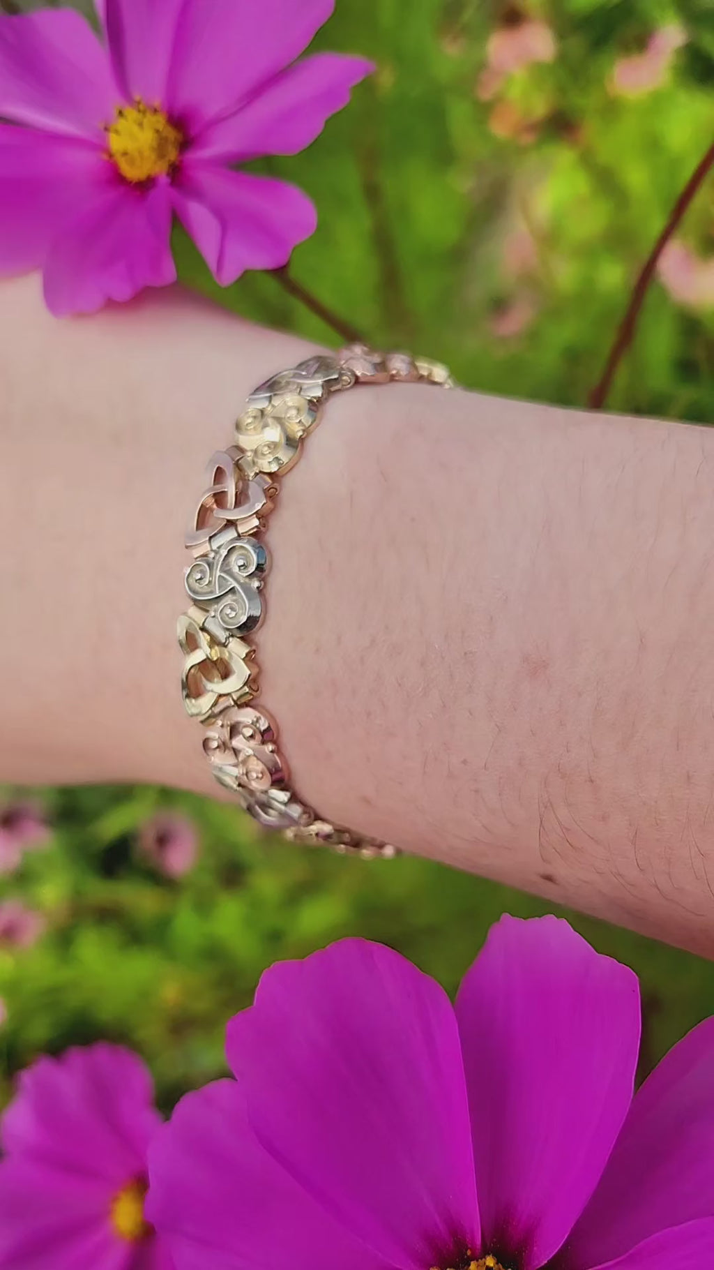 Three Sisters Link Bracelet in tri-color 14K gold worn on the wrist, shown outdoors among cosmos flowers.