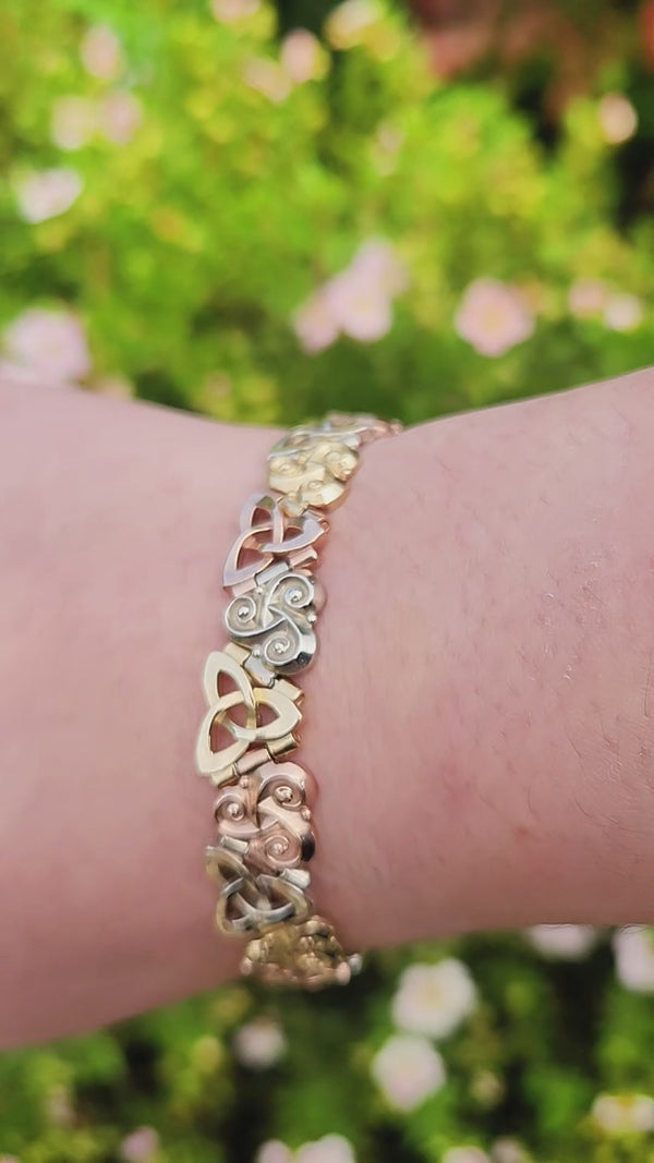 Three Sisters Link Bracelet in tri-color 14K gold worn on the wrist, shown outdoors against green foliage and white blossoms.
